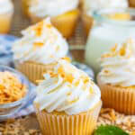 A Coconut Cupcake with tall swirled coconut frosting and toasted coconut in front of a jar of milk.