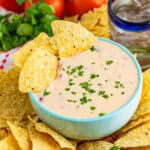 A bowl of creamy queso dip with a tortilla chip scooping it up, garnished with diced tomatoes.