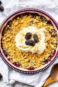 A baking dish of blackberry crisp topped with a scoop of whipped cream.