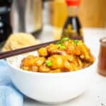 A white bowl of sticky slow cooker chicken teriyaki served over fluffy white rice and garnished with sliced green onions and toasted sesame seeds.