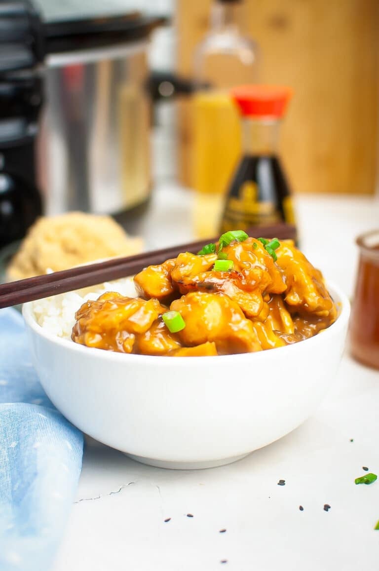 A white bowl of sticky slow cooker chicken teriyaki served over fluffy white rice and garnished with sliced green onions and toasted sesame seeds.