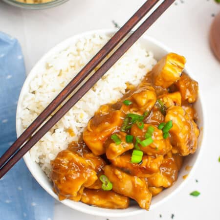 A bowl of slow cooker chicken teriyaki served over white rice with chopsticks resting on the rim and sesame seeds on top.