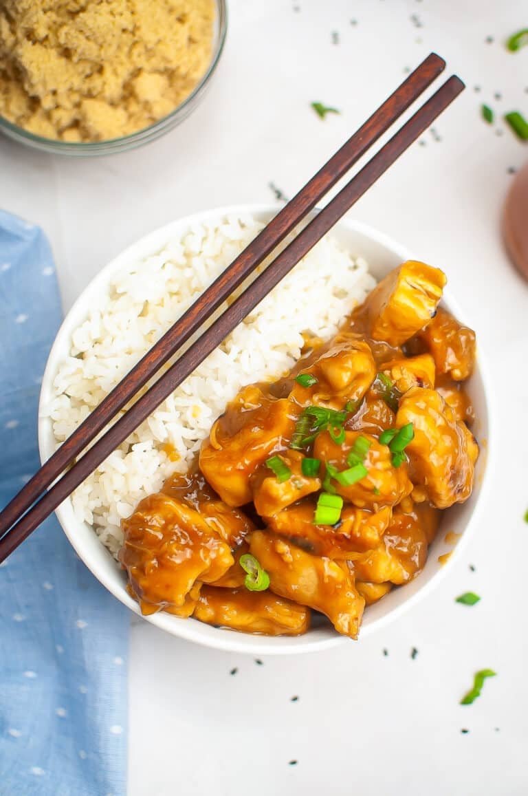 A bowl of slow cooker chicken teriyaki served over white rice with chopsticks resting on the rim and sesame seeds on top.