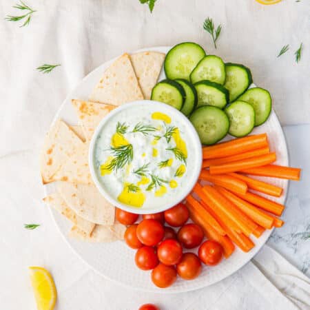 A white bowl of creamy tzatziki sauce drizzled with olive oil and dill, served on a platter with pita, cucumber, carrots, and cherry tomatoes.