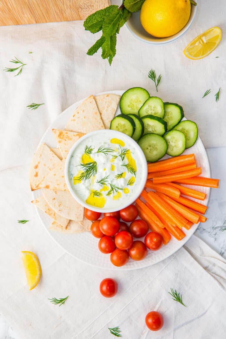 A white bowl of creamy tzatziki sauce drizzled with olive oil and dill, served on a platter with pita, cucumber, carrots, and cherry tomatoes.