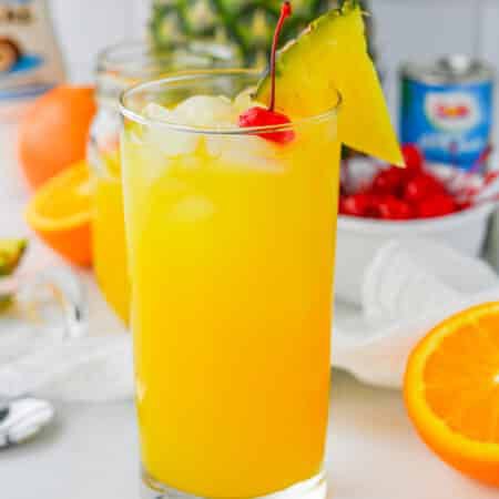 A tall painkiller cocktail over ice garnished with a pineapple wedge and maraschino cherry with a fresh pineapple behind it.