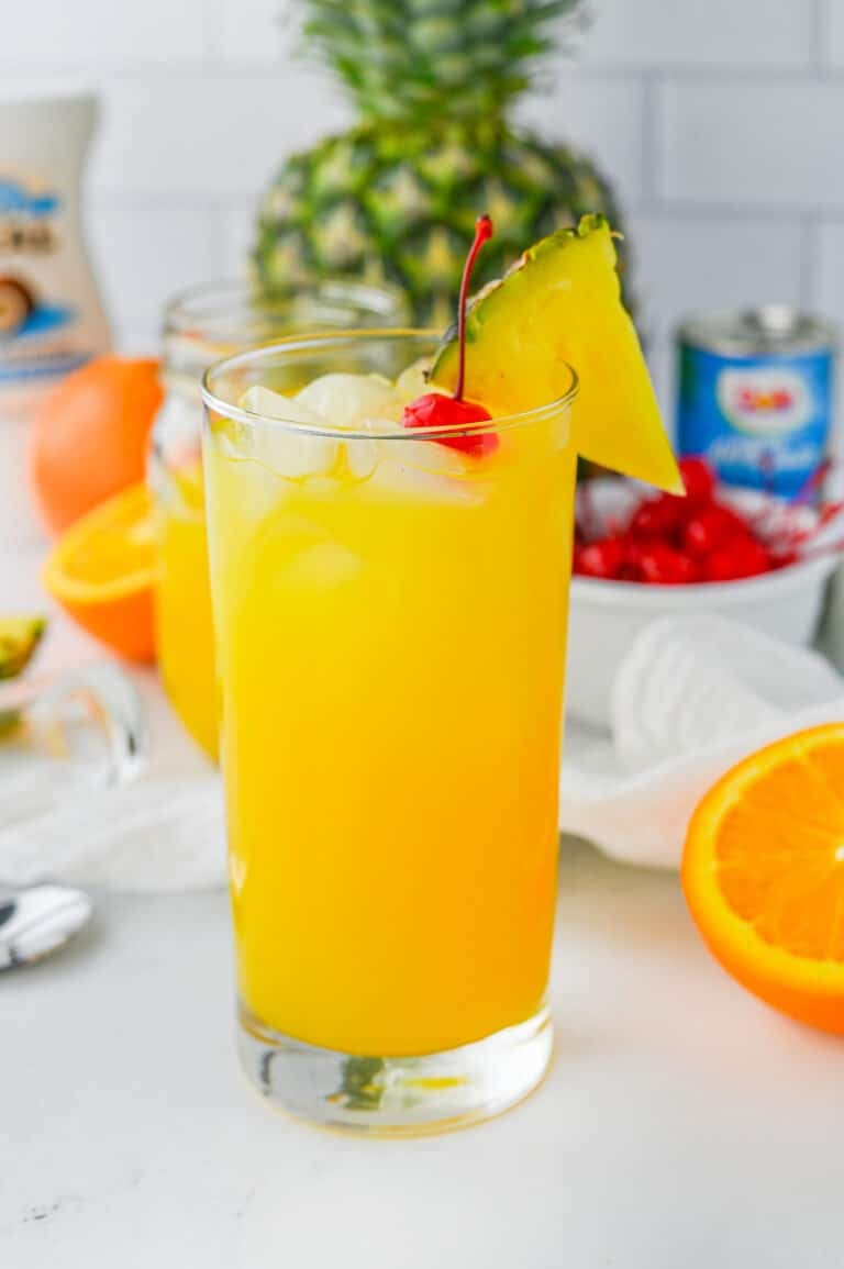 A tall painkiller cocktail over ice garnished with a pineapple wedge and maraschino cherry with a fresh pineapple behind it.