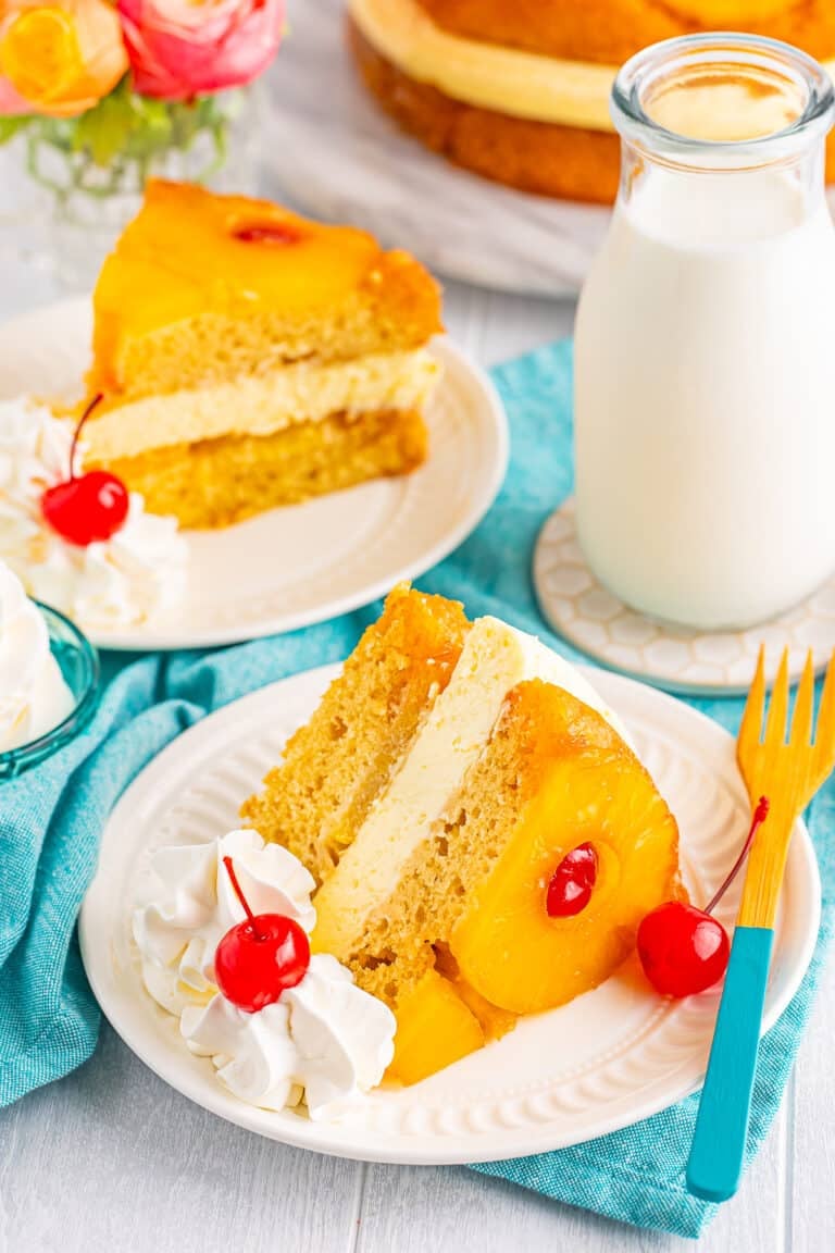 Slice of Copycat Cheesecake Factory Pineapple Upside Down Cheesecake on a white plate showing buttery cake layers, creamy cheesecake middle, and pineapple cherry topping with a glass milk bottle in the background.