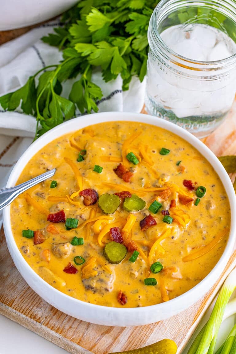 A white bowl of creamy cheeseburger soup topped with crispy bacon, scallions, and cheddar cheese