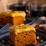 A slice of Pumpkin Cake with praline frosting on a dark plate with a cup of coffee behind it.