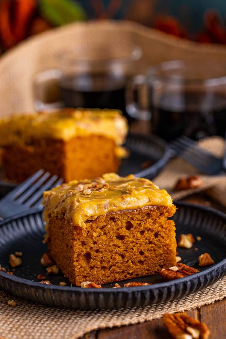 A slice of Pumpkin Cake with praline frosting on a dark plate with a cup of coffee behind it.