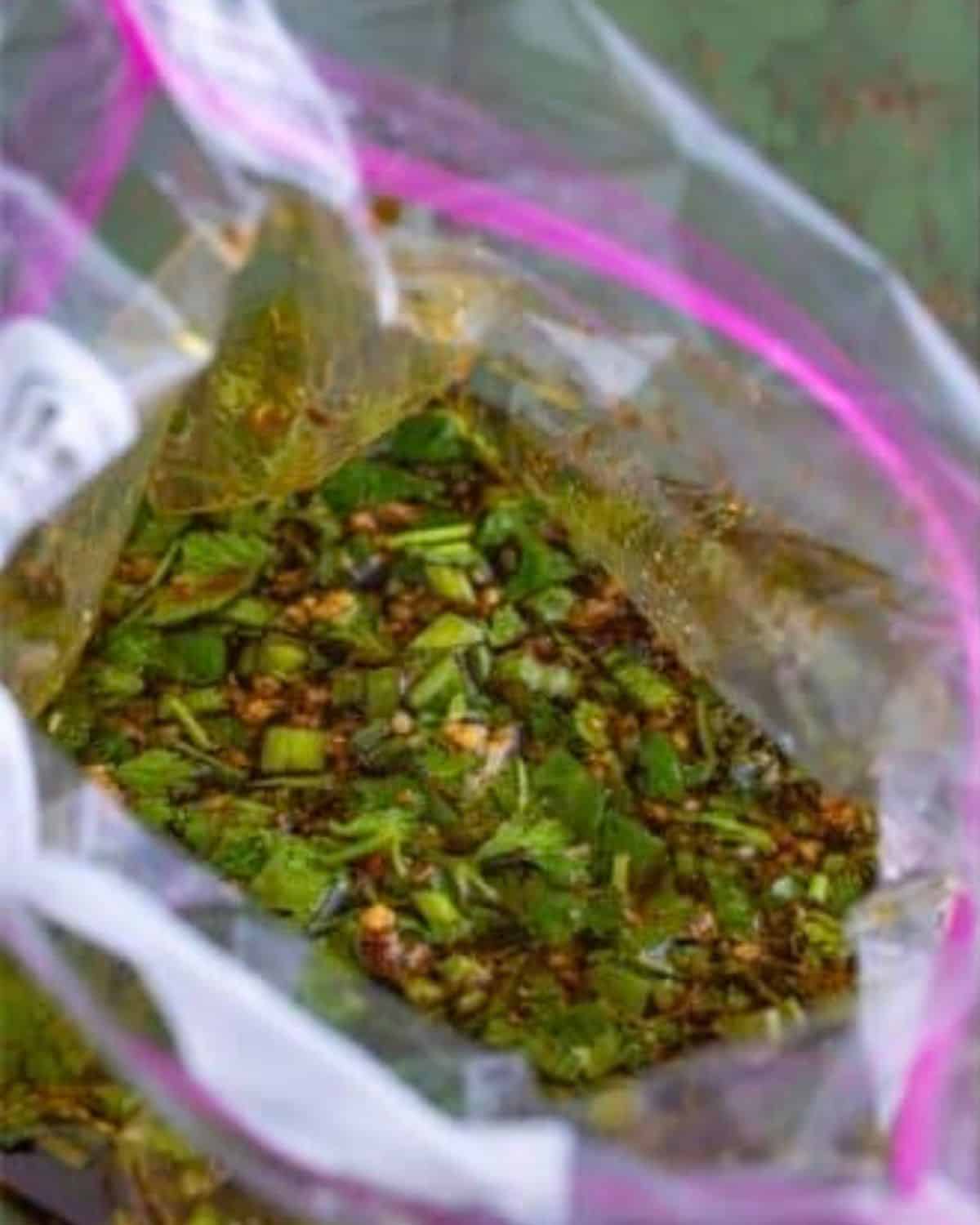 A close-up of a clear resealable plastic bag filled with a marinade containing chopped green herbs, spices, and liquid.