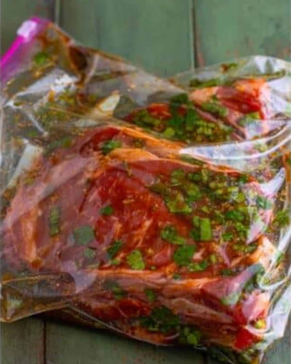 Raw marinated meat with herbs and spices in a sealed plastic ziplock bag, resting on a rustic green wooden surface.