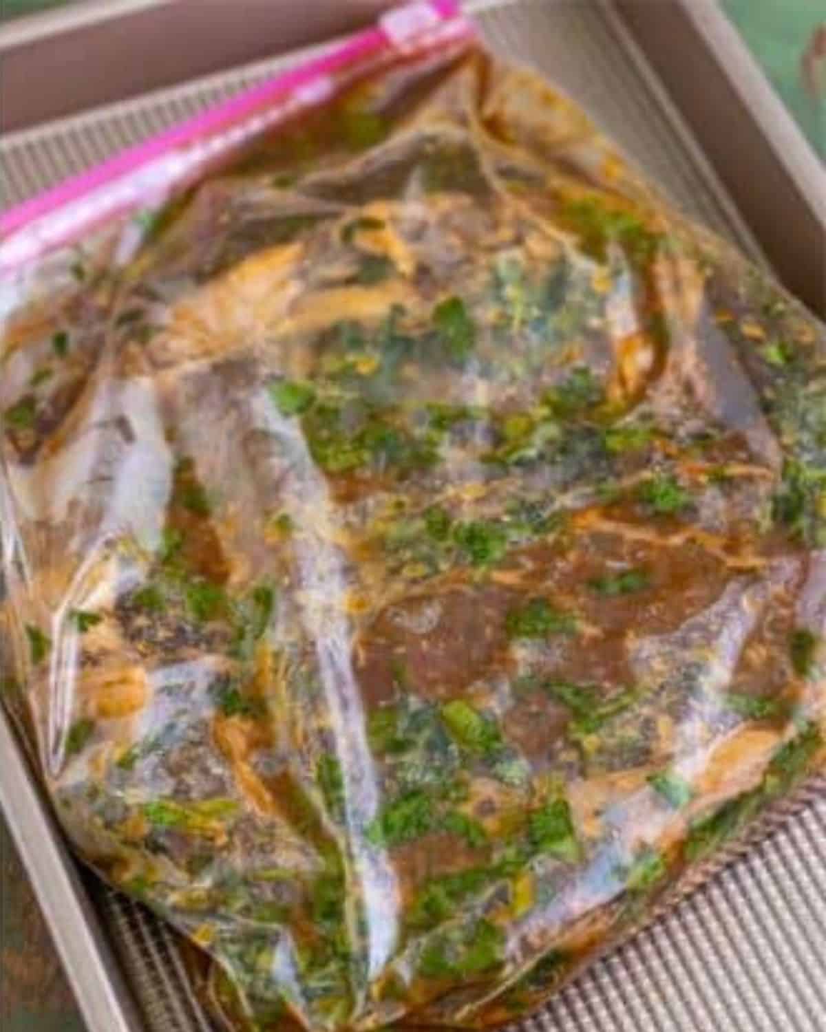 A sealed plastic zip-top bag filled with marinating meat and herbs, placed on a metal baking tray. The marinade is dark with visible green herbs.