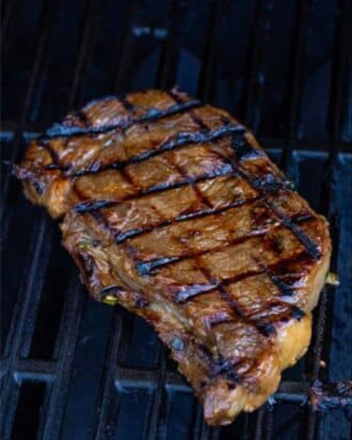 A grilled steak with char marks sits on a barbecue grill, displaying a golden-brown, seared surface with dark grill lines.