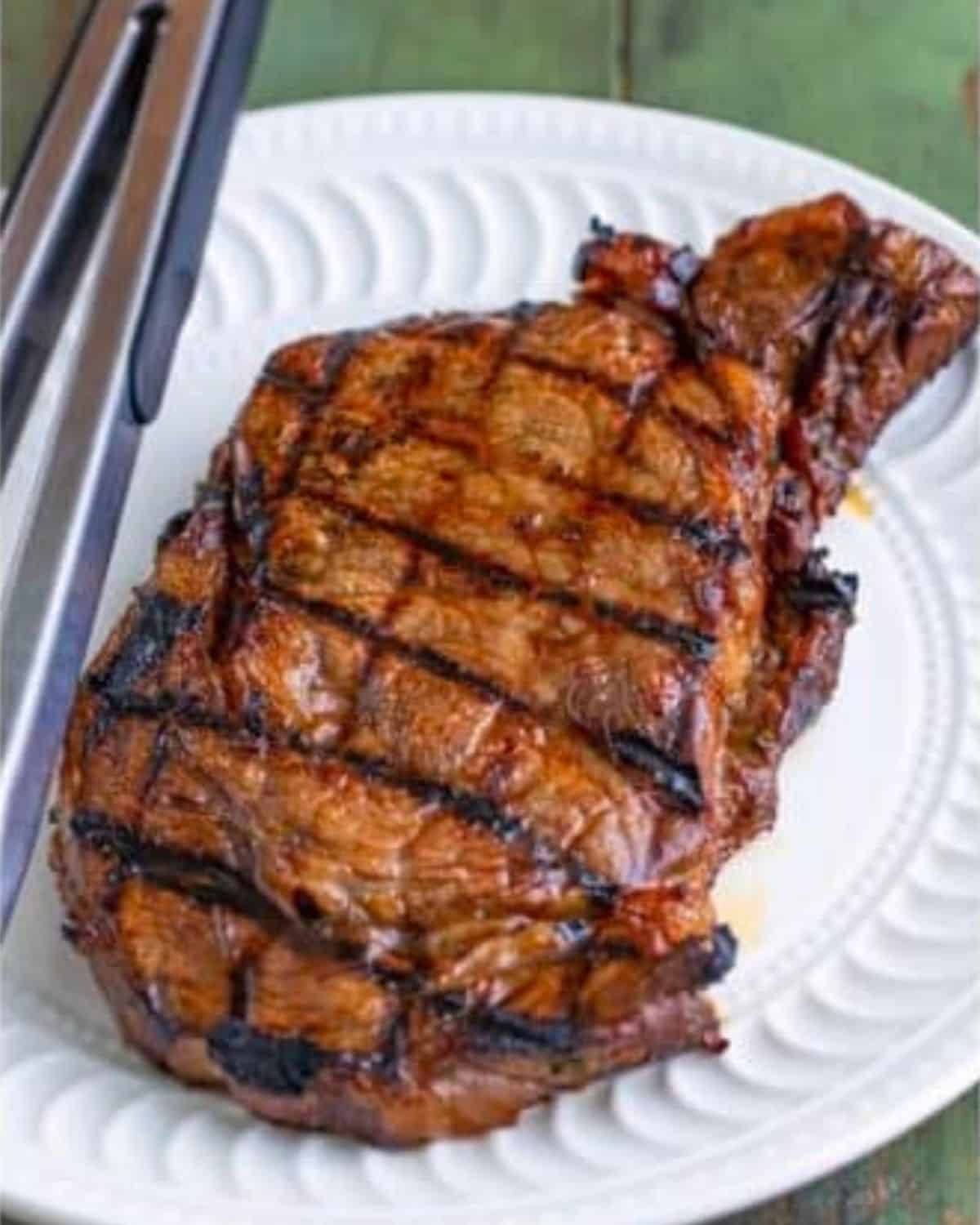 A grilled steak with visible char marks sits on a white plate, next to a pair of metal tongs on a green surface.