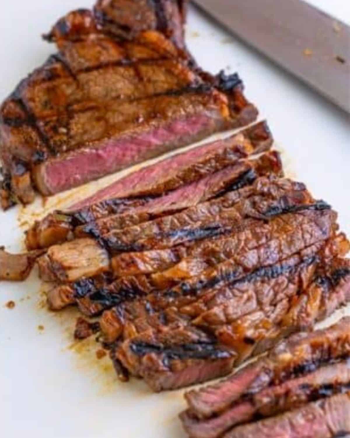 Sliced grilled steak with visible grill marks and a juicy, medium-rare interior on a white cutting board, next to a large knife.