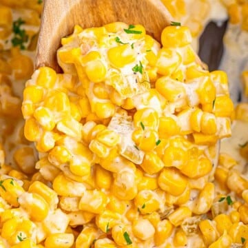 A wooden spoon lifting a scoop of creamy slow cooker creamed corn with green chiles over the crock.