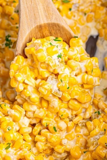 A wooden spoon lifting a scoop of creamy slow cooker creamed corn with green chiles over the crock.