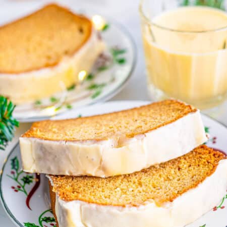Thick slices of glazed eggnog bread stacked on a holiday plate with a glass of eggnog behind.
