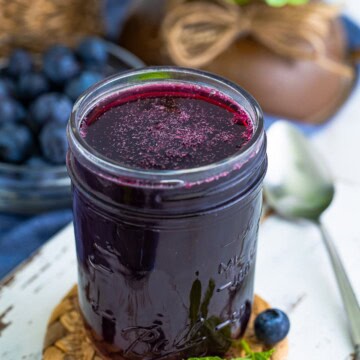 A mason jar of deep purple Blueberry Syrup with fresh blueberries and mint on a white board.