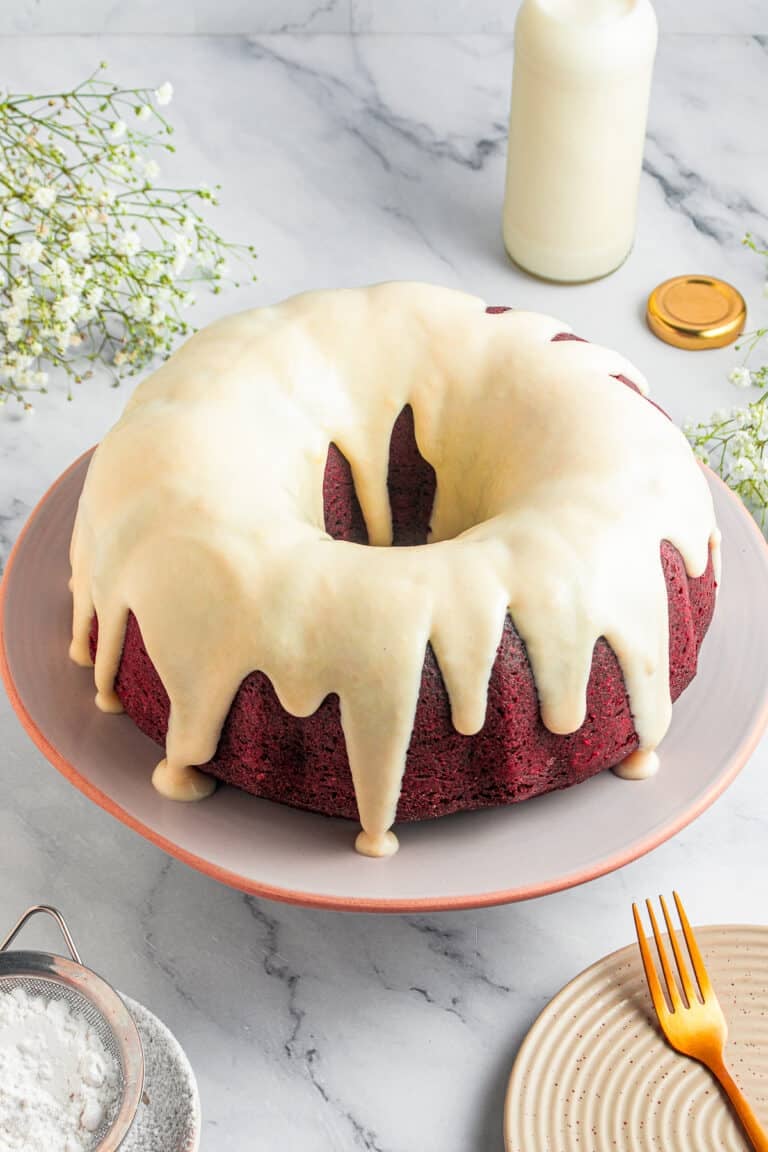 A whole red velvet bundt cake with cream cheese glaze dripping down the sides on a pink stand.