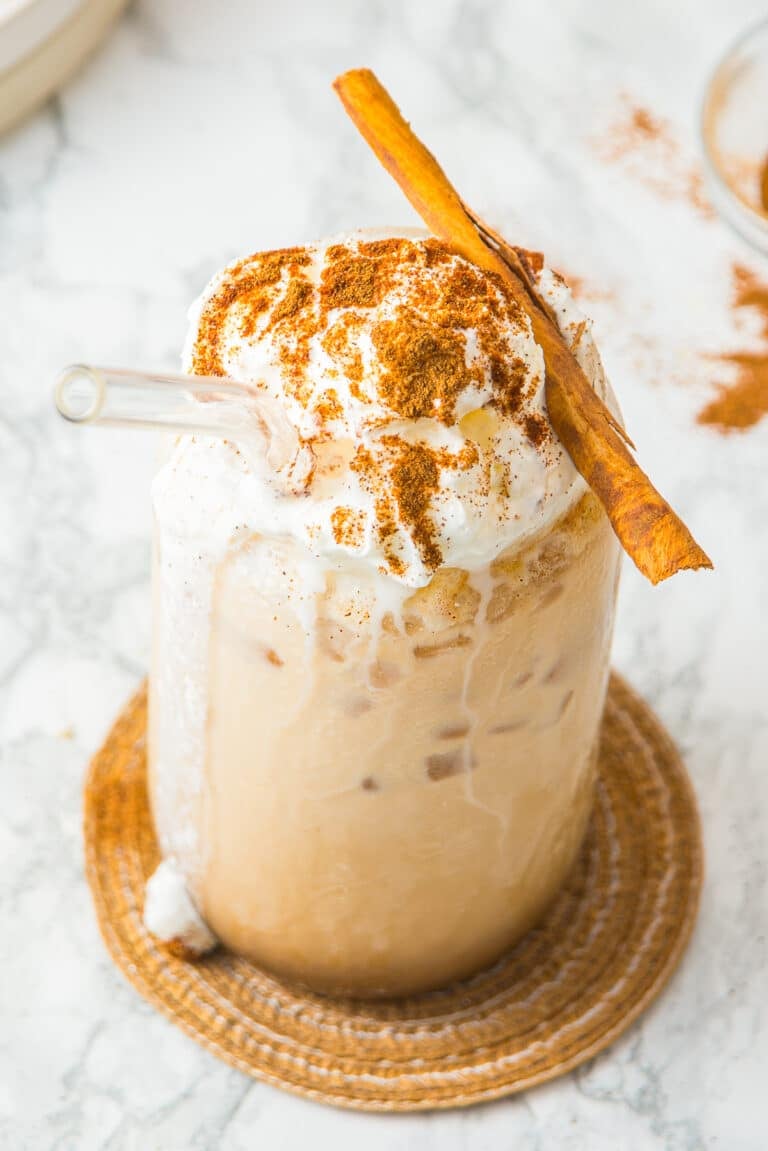 An iced pumpkin spice latte topped with whipped cream and a cinnamon stick on a marble counter.