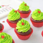 Grinch cupcakes with tall bright green frosting swirls and red heart sprinkles on a holiday towel.