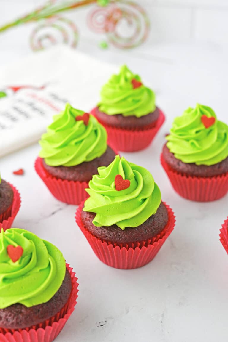Grinch cupcakes with tall bright green frosting swirls and red heart sprinkles on a holiday towel.
