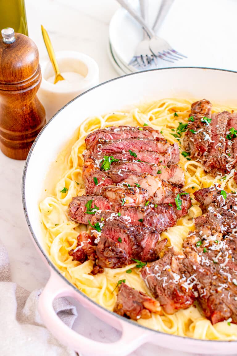 Steak Alfredo in a skillet with sliced ribeye over creamy fettuccine garnished with Parmesan and parsley.