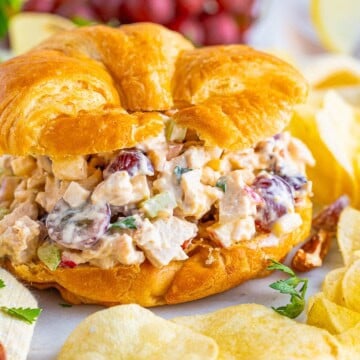 Chicken salad piled high on a flaky croissant with chips, grapes, and apple slices on the side.