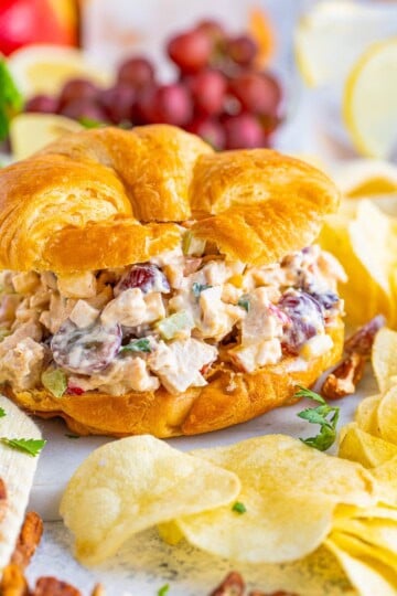 Chicken salad piled high on a flaky croissant with chips, grapes, and apple slices on the side.