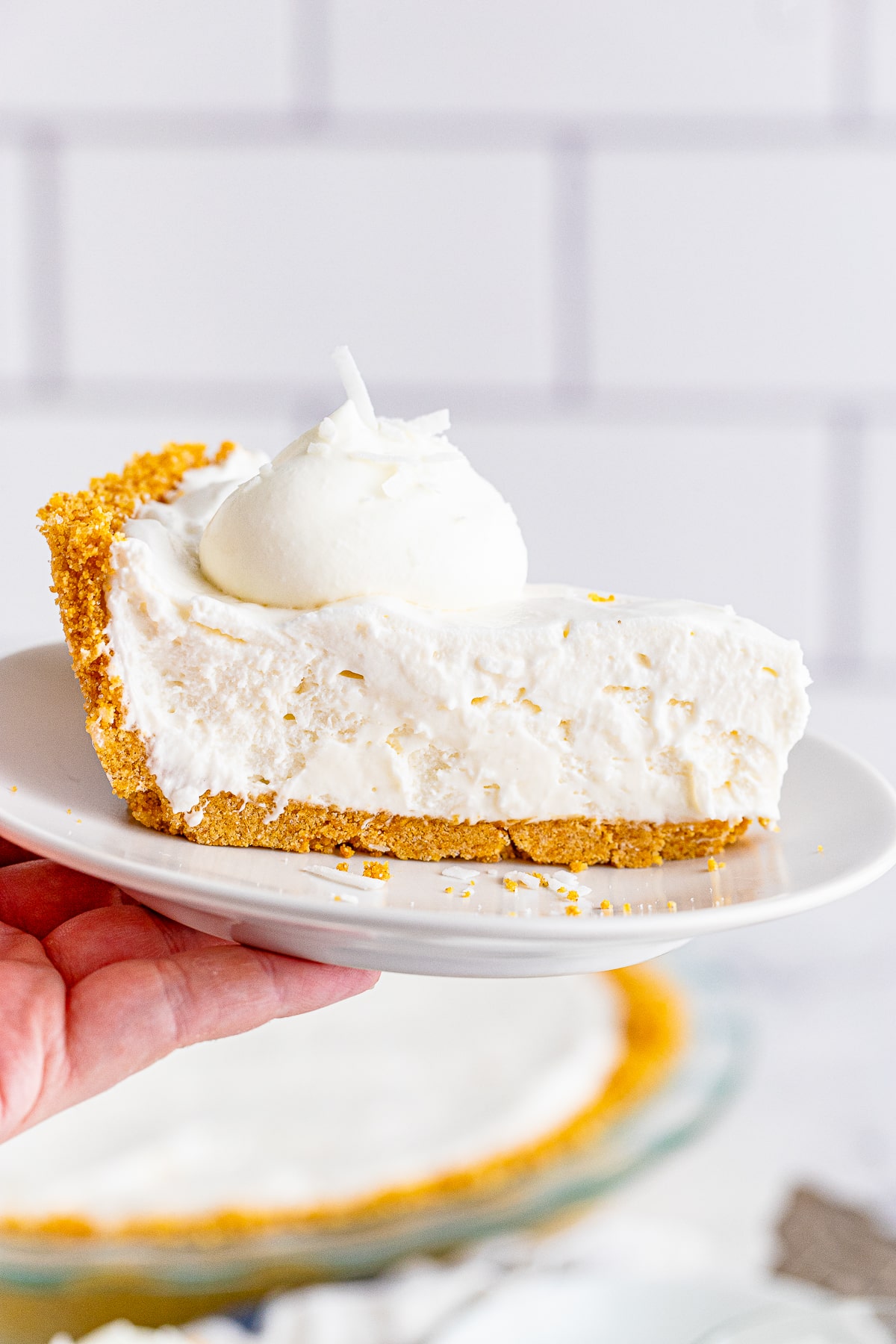 a hand holding up a slice of coconut cream pie on a plate in air