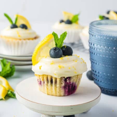 A Lemon Blueberry Cupcake on a small stand topped with cream cheese frosting, blueberries, lemon, and mint.