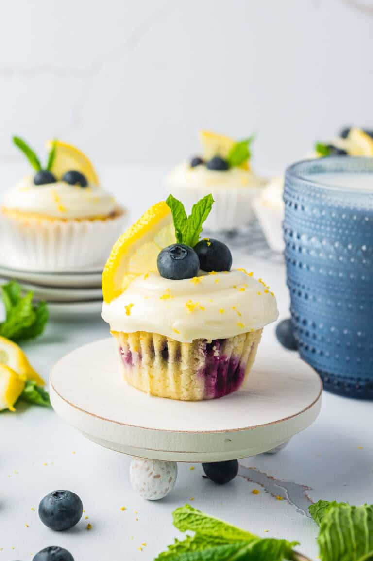 A Lemon Blueberry Cupcake on a small stand topped with cream cheese frosting, blueberries, lemon, and mint.