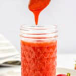 A spoon drizzling thick homemade Strawberry Sauce into a glass jar.