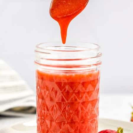 A spoon drizzling thick homemade Strawberry Sauce into a glass jar.