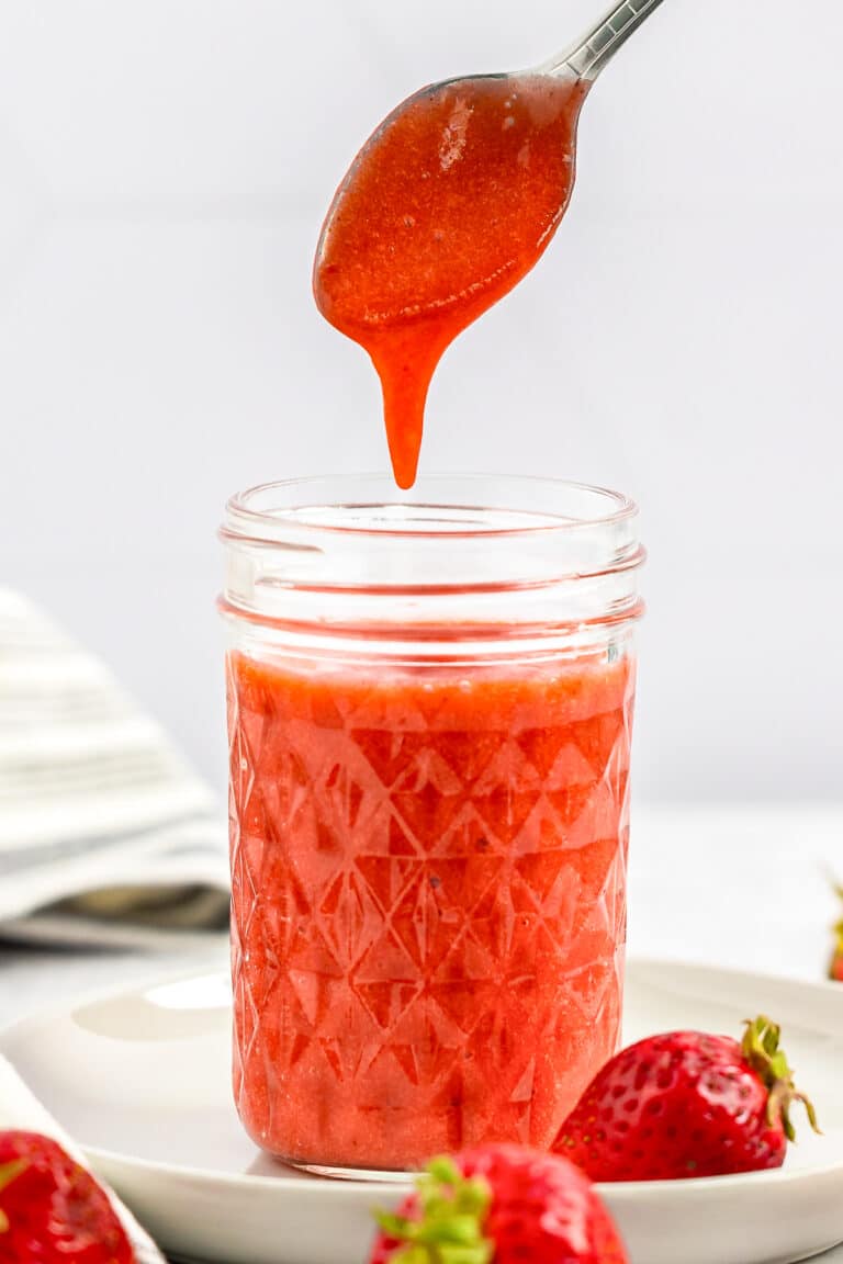 A spoon drizzling thick homemade Strawberry Sauce into a glass jar.