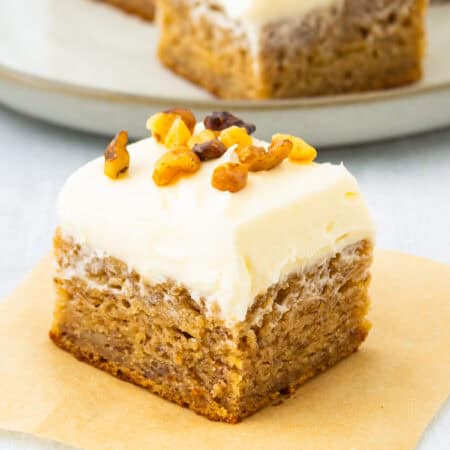Frosted banana bars stacked on parchment paper with cream cheese frosting and walnuts.