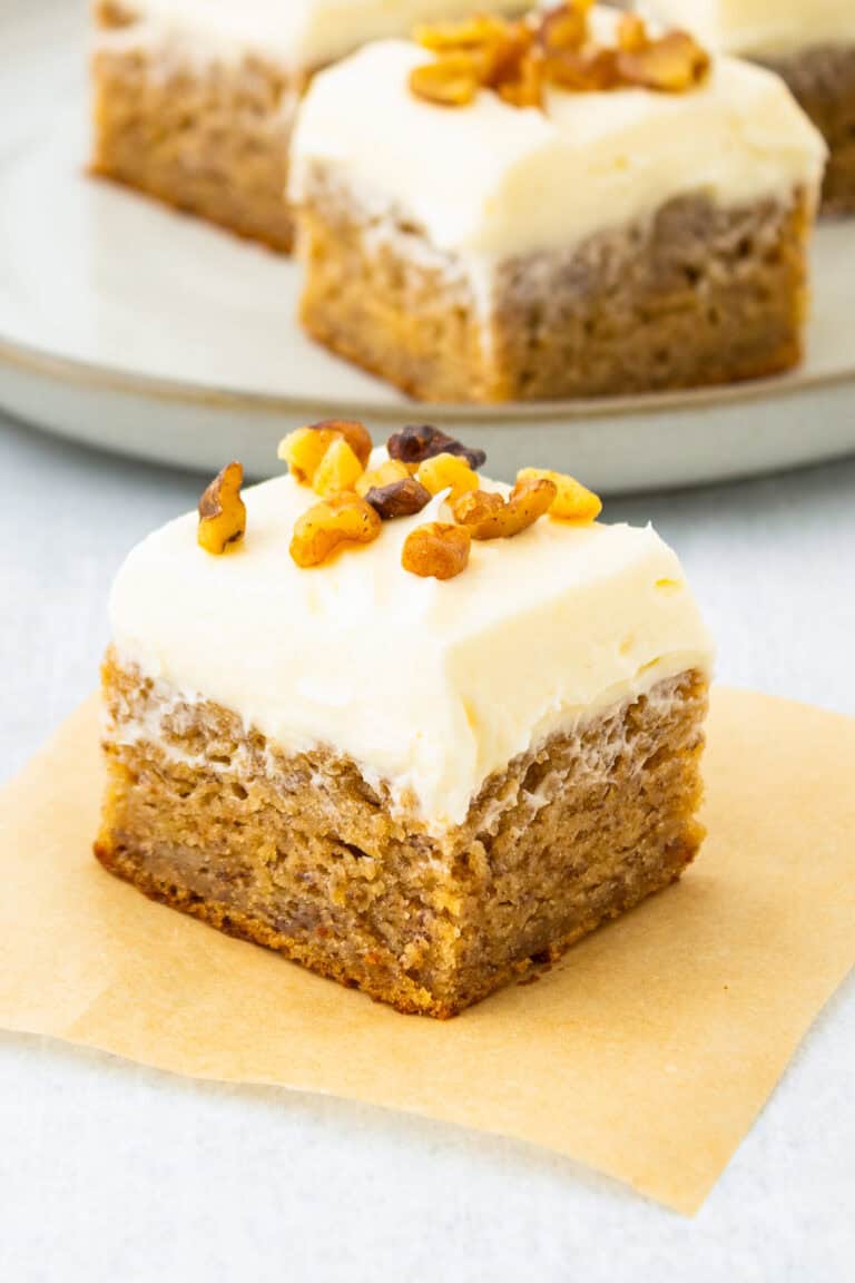 Frosted banana bars stacked on parchment paper with cream cheese frosting and walnuts.