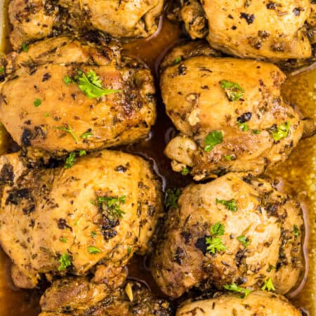 Close-up of golden baked chicken thighs stacked on a plate, glazed with lemon-garlic pan sauce and fresh parsley.