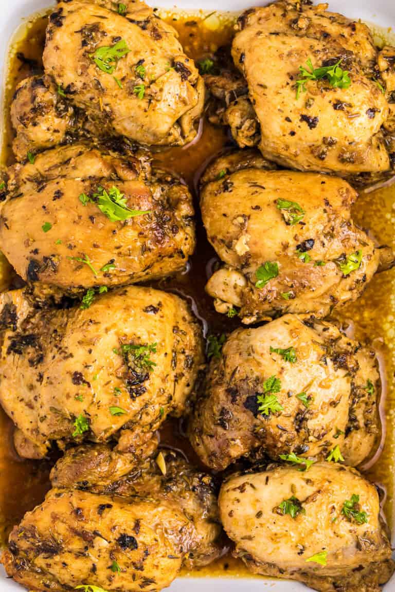 Close-up of golden baked chicken thighs stacked on a plate, glazed with lemon-garlic pan sauce and fresh parsley.