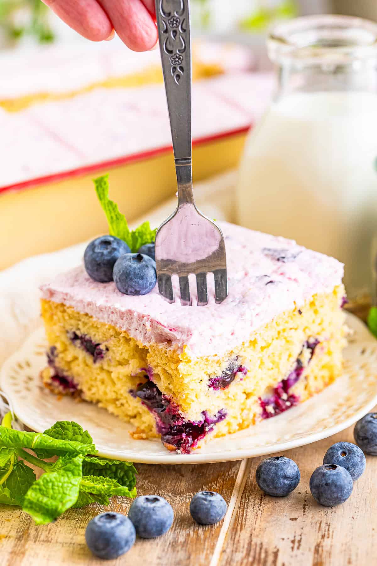 A slice of Blueberry Cake topped with pink blueberry frosting and fresh blueberries.