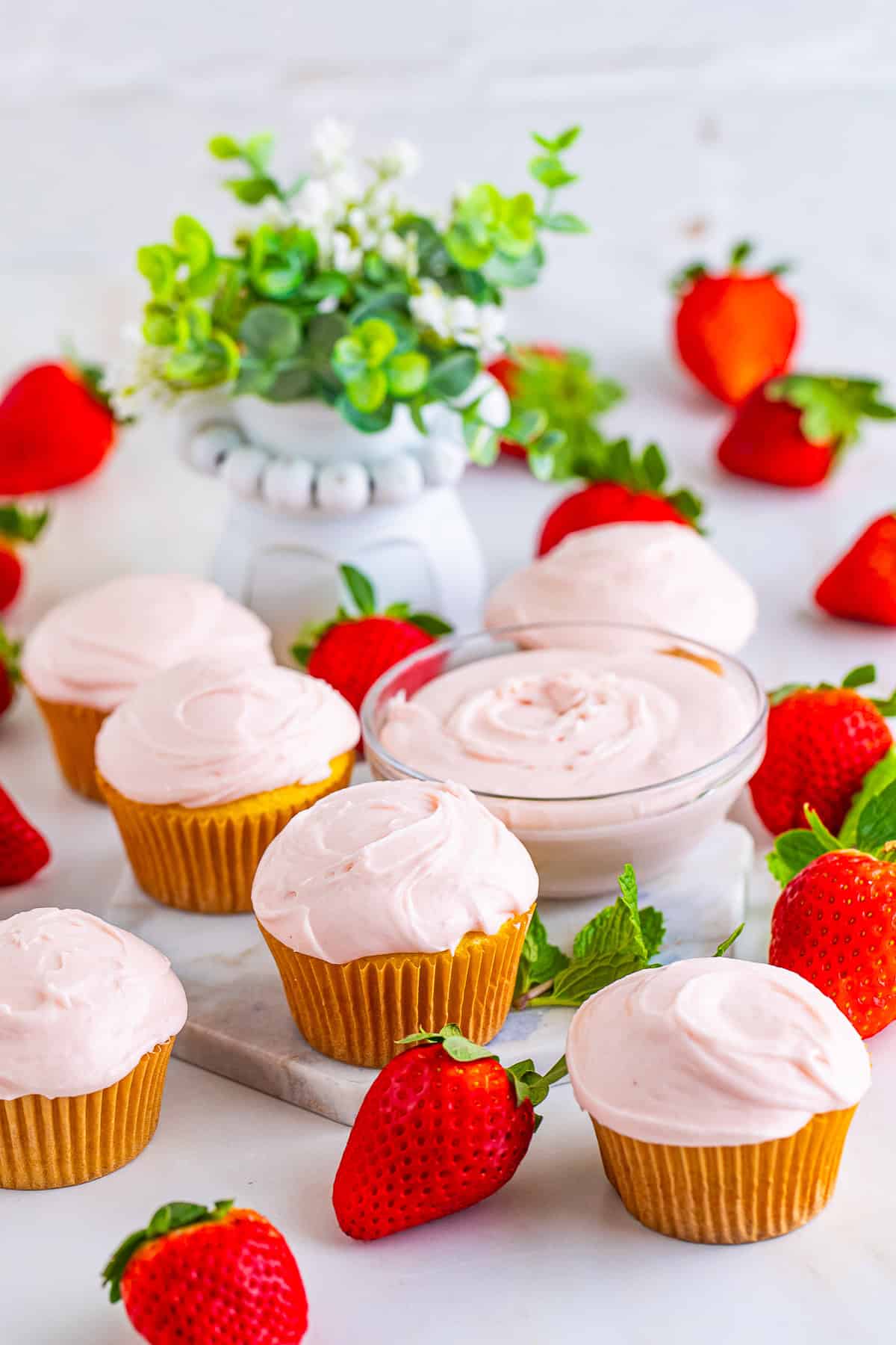 A bowl of fresh strawberry cream cheese frosting surrounded by frosted cupcakes and strawberries.
