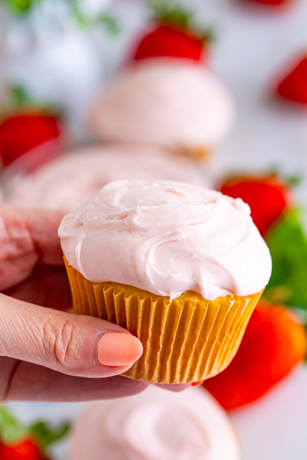 a hand holding up a cupcake topped with Strawberry Cream Cheese Frosting in air