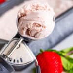 A scoop of homemade Strawberry Ice Cream in an ice cream scooper resting on a frozen pan of ice cream with fresh strawberries.