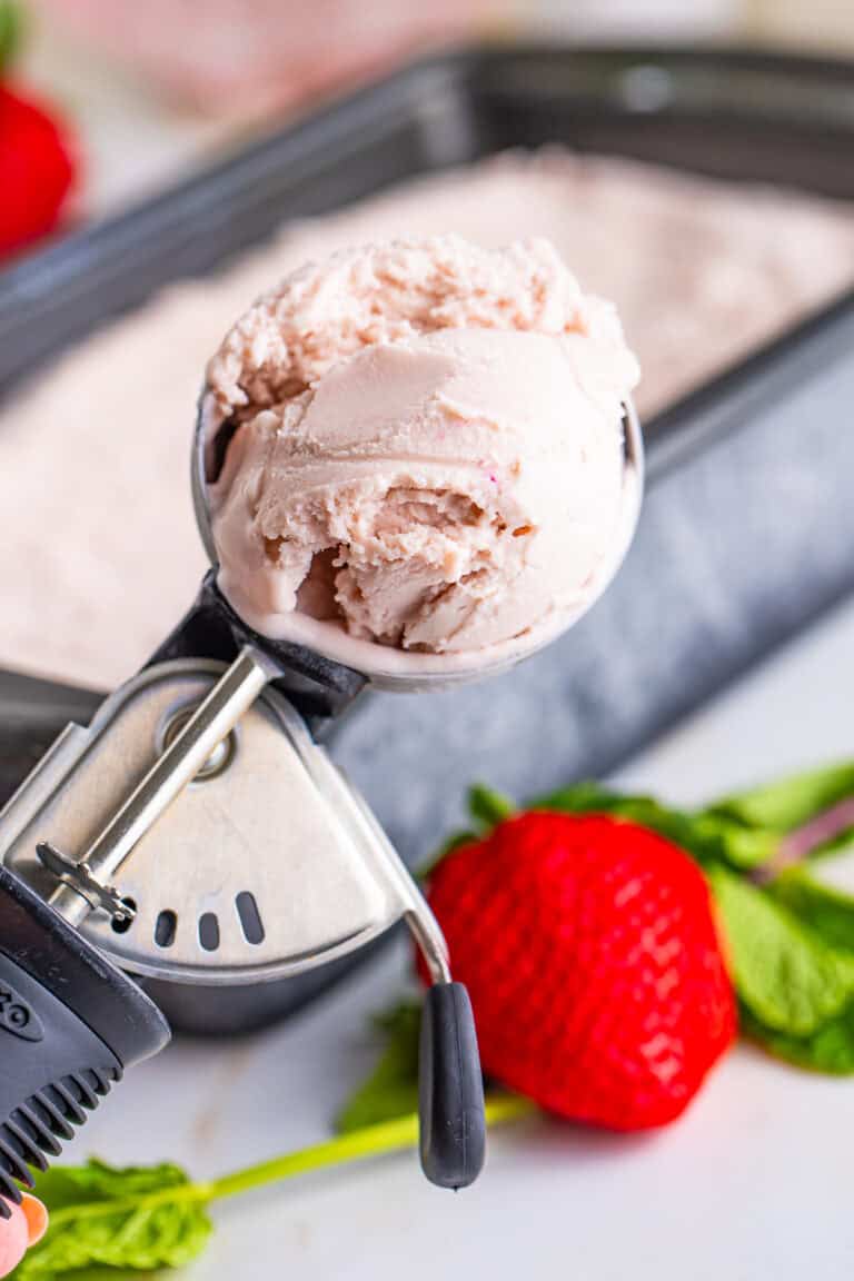 A scoop of homemade Strawberry Ice Cream in an ice cream scooper resting on a frozen pan of ice cream with fresh strawberries.