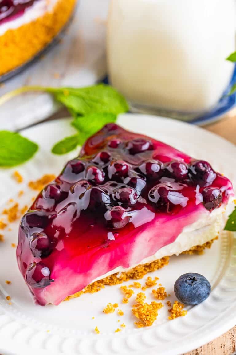 A slice of No Bake Blueberry Cheesecake on a white plate with glossy blueberry topping over the creamy filling and graham crust.