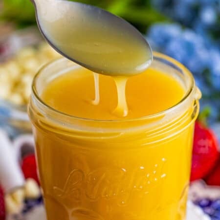 A spoon drizzling thick White Chocolate Sauce back into a glass jar.