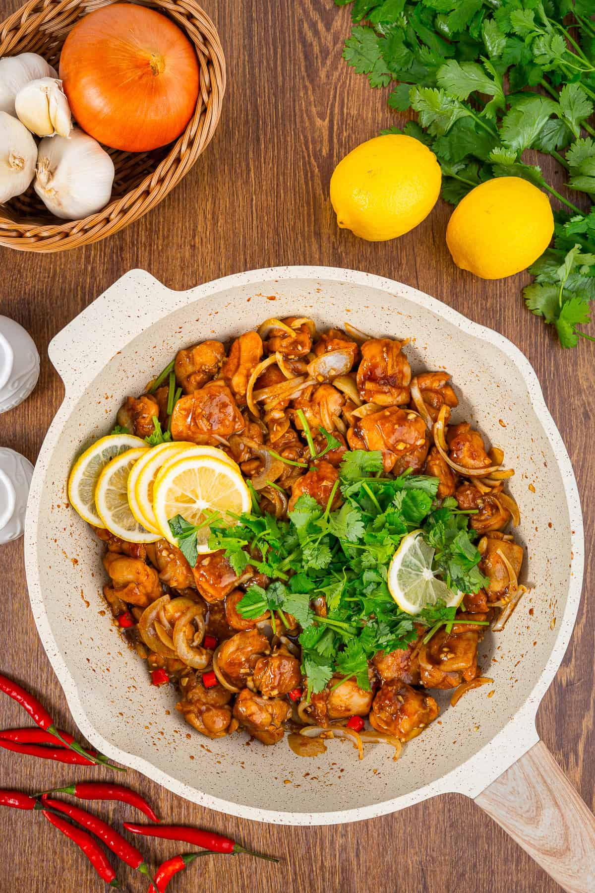 Asian lemon chicken in a pan garnished with cilantro and lemon, surrounded by garlic, onion, and fresh lemons.